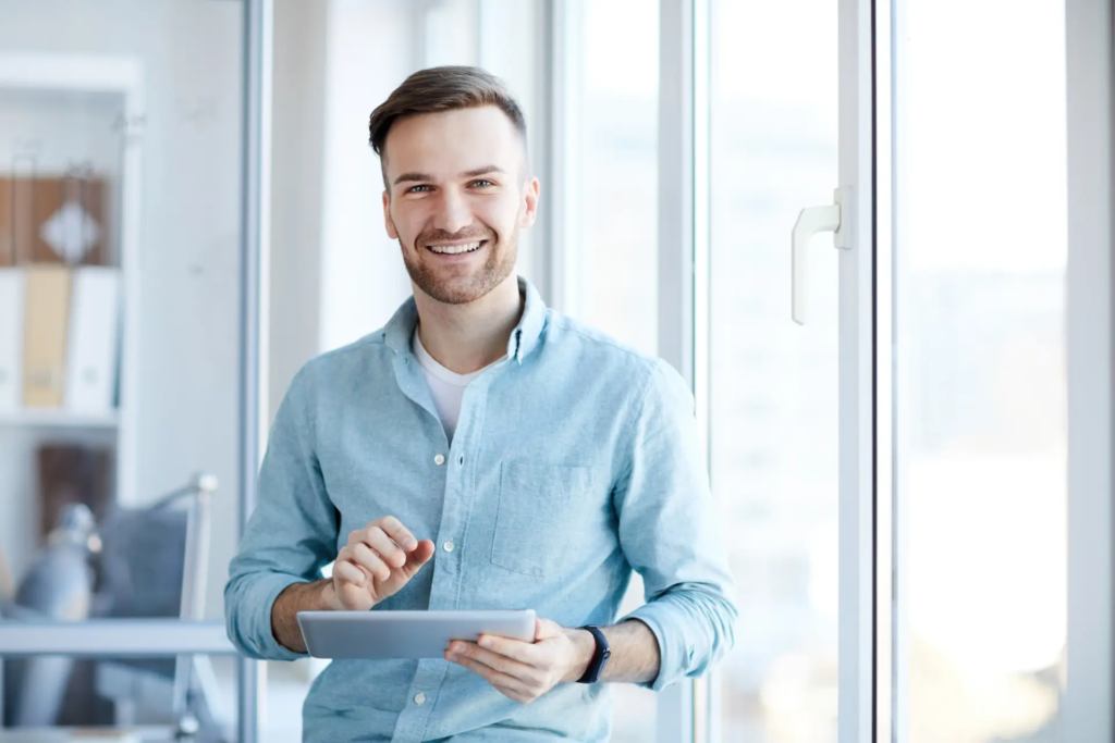  Un jeune homme souriant, vêtu d'une chemise claire, tient une tablette numérique et regarde la caméra. Il se trouve dans un bureau lumineux avec de grandes fenêtres en arrière-plan.