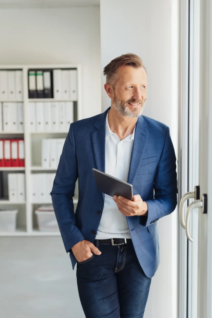 Un homme d'affaires souriant et barbu, vêtu d'une veste de costume bleue et d'un jean, tient une tablette numérique. Il regarde par une fenêtre, avec des étagères remplies de dossiers en arrière-plan dans un bureau lumineux.
