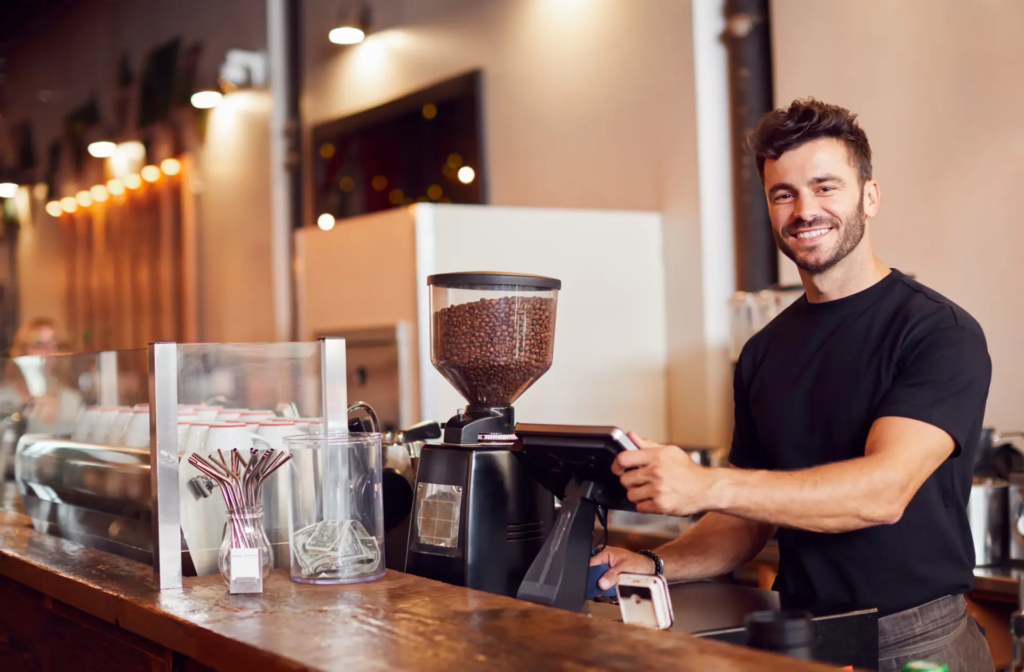 Un barista souriant et barbu, vêtu d'un t-shirt noir, est debout derrière le comptoir d'un café, manipulant un moulin à café ou une tablette. L'arrière-plan flou montre l'intérieur moderne du café.