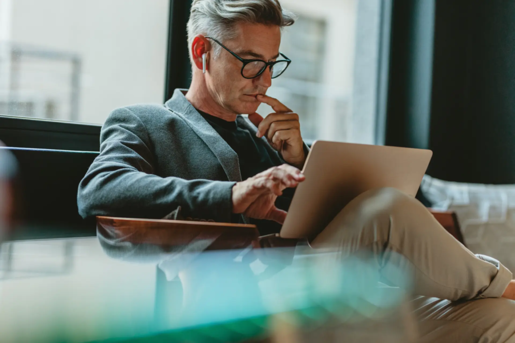 Un homme d'affaires d'âge mûr, portant des lunettes et des écouteurs sans fil, est assis et concentré sur son ordinateur portable. Il semble travailler ou réfléchir intensément, le doigt sur le menton.