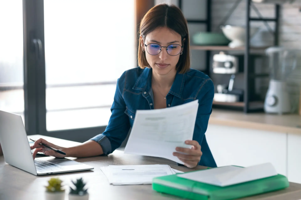 Une jeune femme portant des lunettes est assise à une table, travaillant sur un ordinateur portable et examinant des documents. La scène suggère du télétravail ou la gestion de papiers personnels dans un intérieur lumineux.
