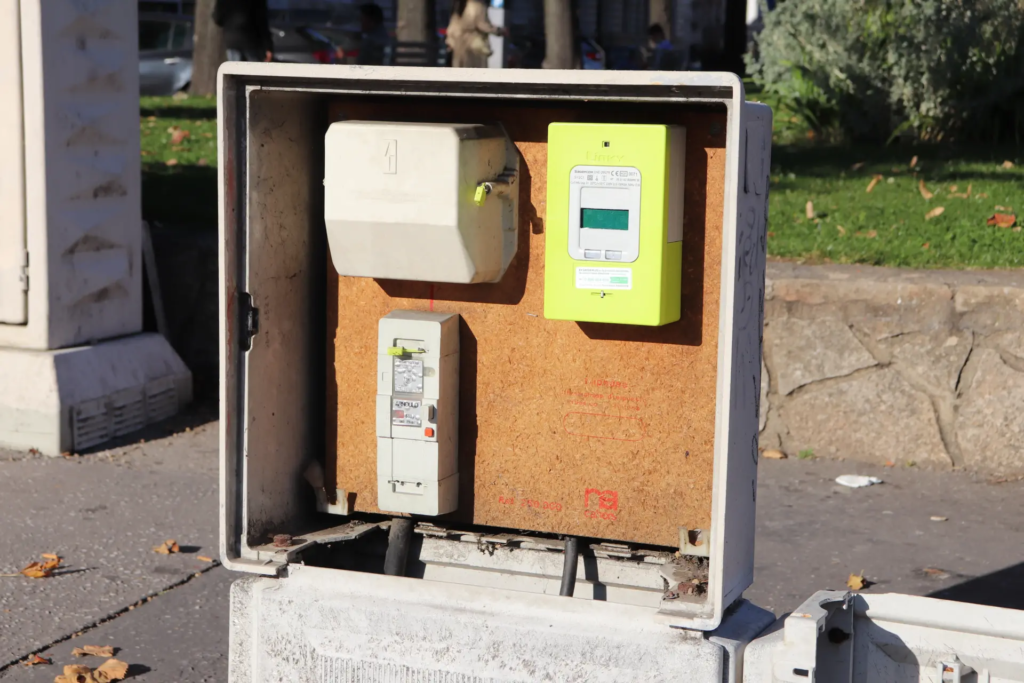 Un boîtier de raccordement extérieur ouvert révélant un compteur électrique Linky de couleur vert clair et d'autres équipements électriques. L'ensemble est monté sur un panneau en aggloméré, avec un environnement urbain en arrière-plan.