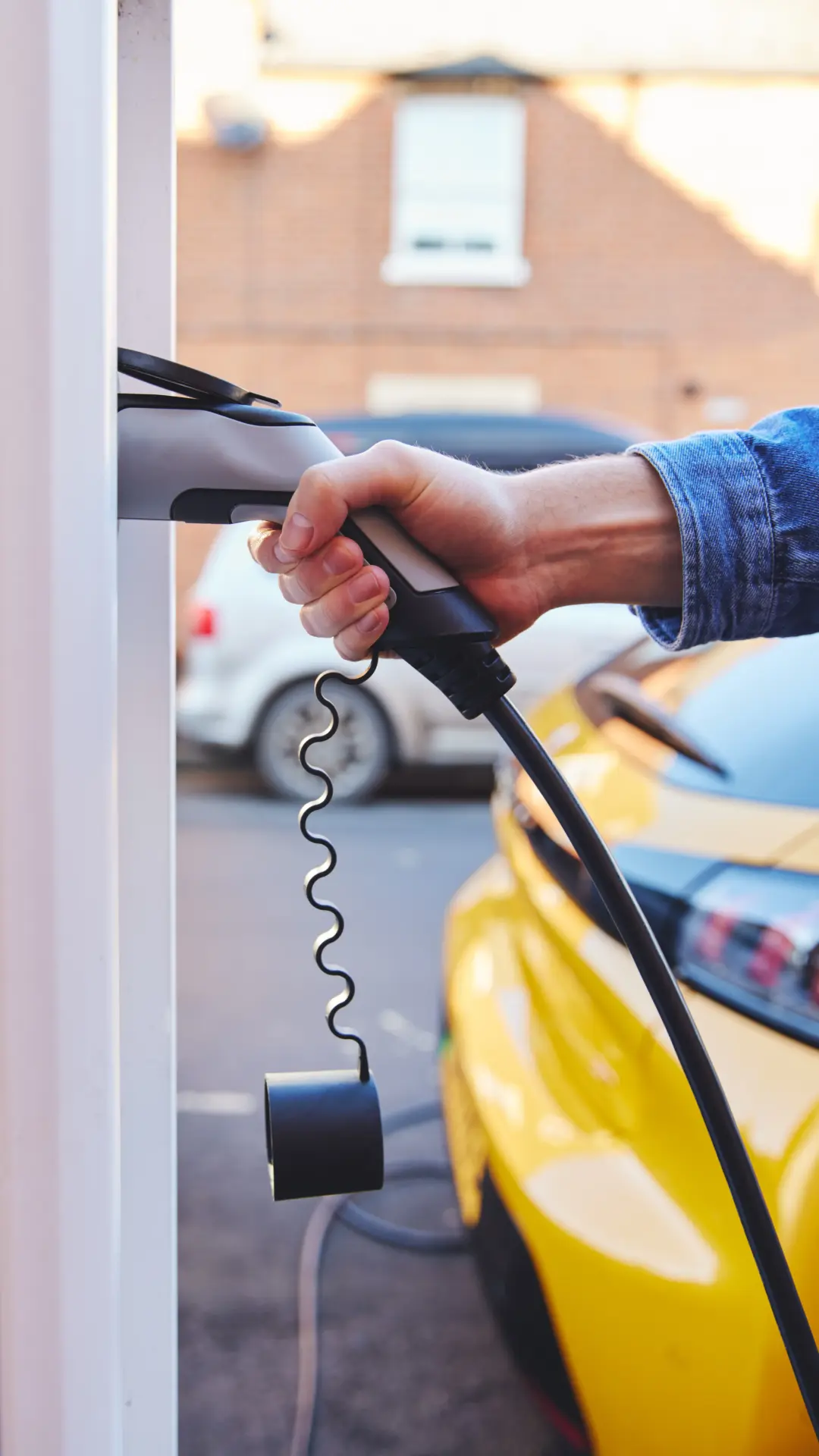 Une main masculine est en train de brancher un câble de recharge sur une borne murale pour voiture électrique. L'avant d'une voiture jaune est visible en arrière-plan, suggérant la recharge d'un véhicule à domicile ou dans un lieu public.