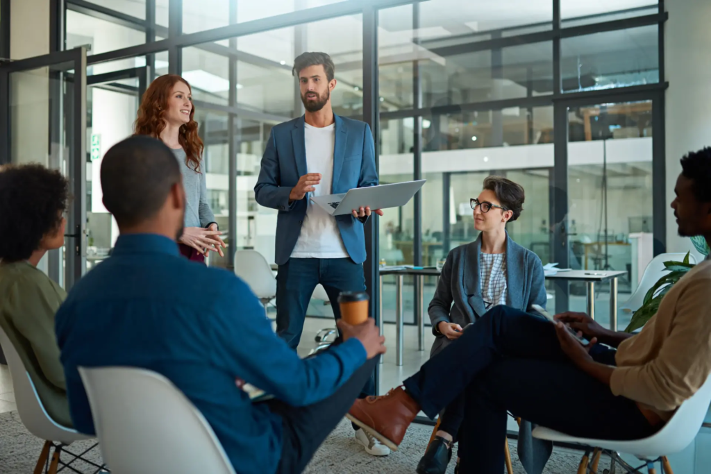 Un homme se tient debout, tenant un ordinateur portable, et s'adresse à un groupe de collègues assis en cercle dans un espace de bureau moderne. La scène illustre une présentation ou une session de brainstorming en équipe.