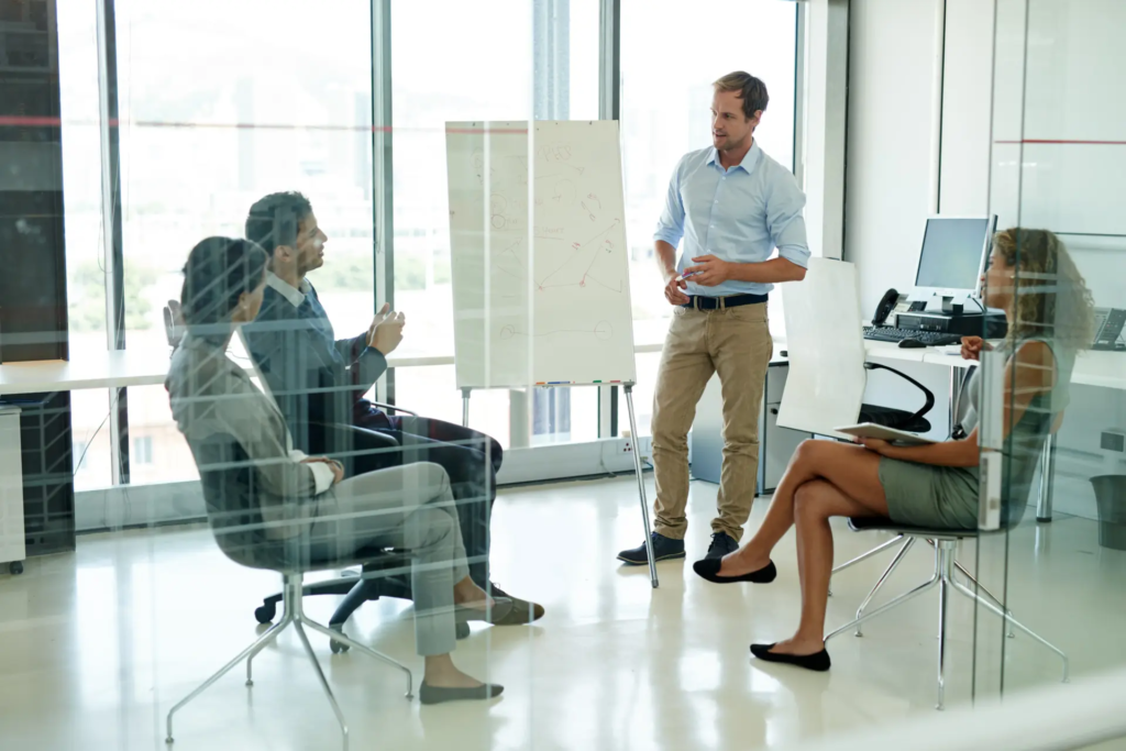 Un homme se tient debout devant un tableau blanc, s'adressant à trois collègues assis dans un bureau lumineux aux grandes baies vitrées. La scène dépeint une réunion informelle, une présentation ou un brainstorming en équipe.