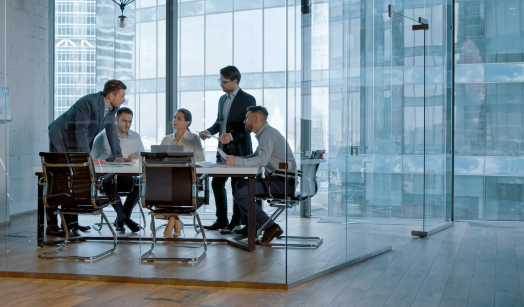 Un groupe de cinq professionnels en tenue de bureau discute autour d'une table dans une salle de réunion vitrée avec une vue sur des gratte-ciel. La scène dépeint une discussion ou une présentation collaborative.