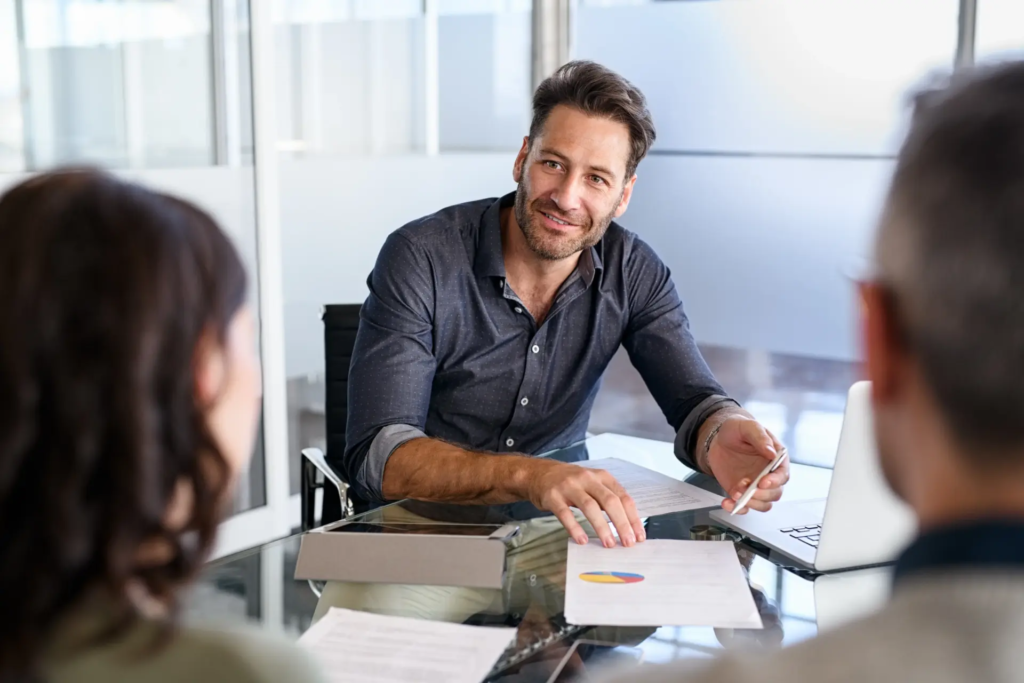 Un homme d'affaires souriant et barbu est assis à une table, regardant deux clients avec des documents et un ordinateur portable devant lui. L'image suggère une consultation, une présentation ou une réunion importante.
