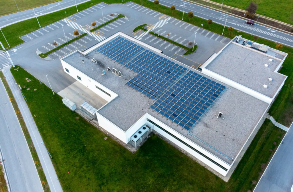 Vue aérienne d'un bâtiment commercial ou industriel avec un grand toit couvert de panneaux solaires bleus. Un vaste parking vide est visible autour du bâtiment, entouré de verdure et de routes.