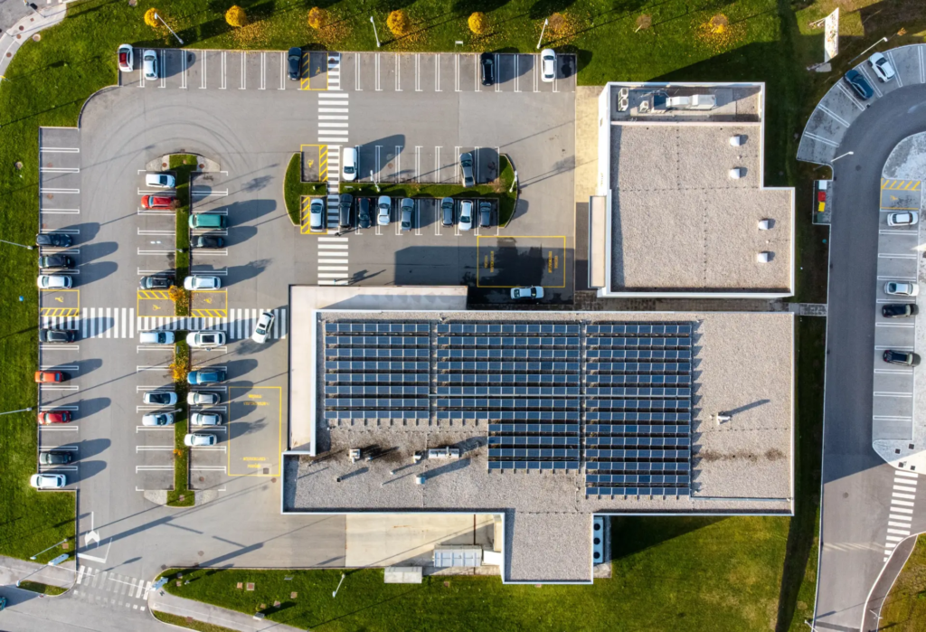 Vue aérienne d'un bâtiment commercial ou industriel dont le toit est couvert de nombreux panneaux solaires, avec un grand parking rempli de voitures à côté. La zone est entourée de pelouses vertes et de routes.