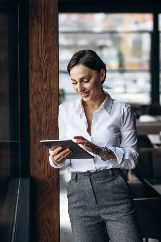 Une femme souriante en chemise blanche et pantalon gris utilise une tablette numérique, appuyée contre un pilier en bois dans un environnement de bureau ou de café. Elle semble engagée dans son travail numérique.