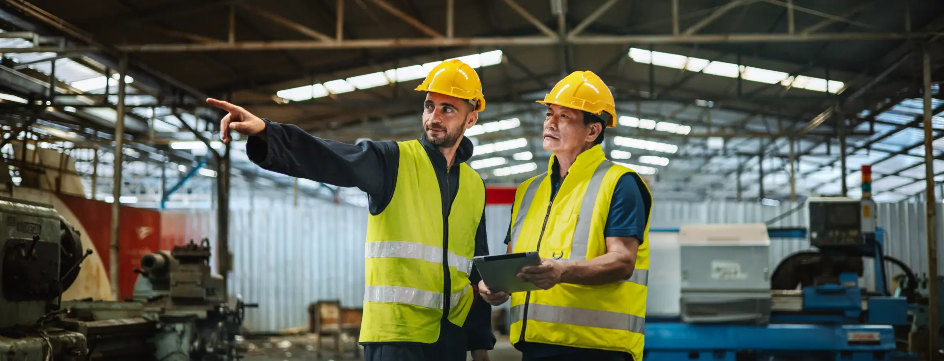 Deux hommes en casques de sécurité jaunes et gilets réfléchissants se tiennent dans un environnement industriel. L'un pointe du doigt tandis que l'autre tient une tablette, suggérant une inspection ou une discussion sur site.