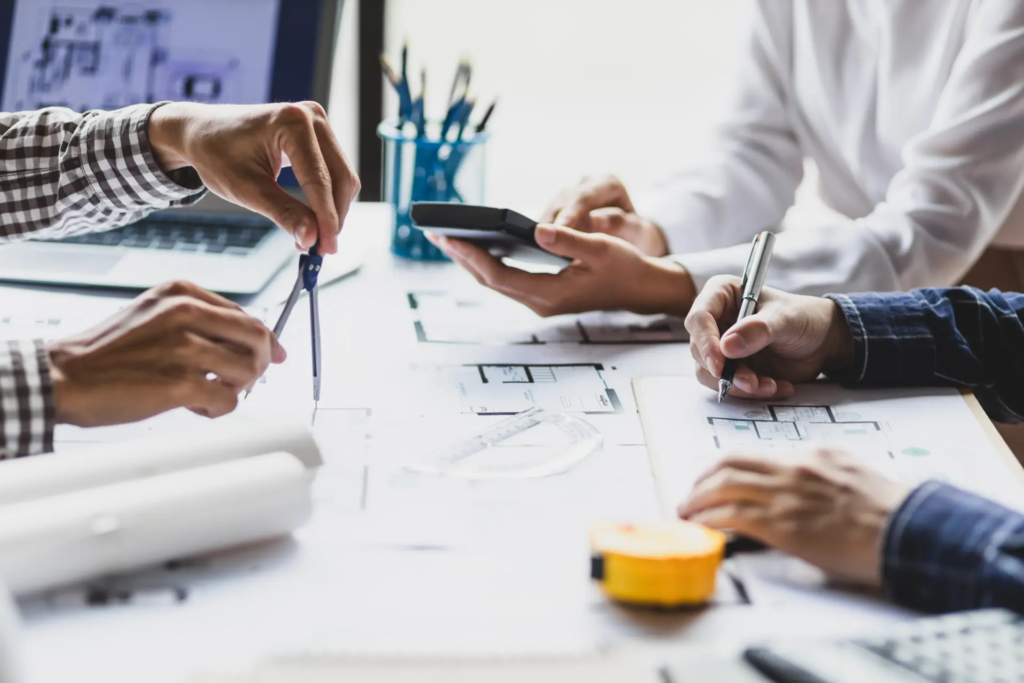 Trois personnes collaborent autour de plans architecturaux et de documents sur une table, l'une tenant un compas, une autre un stylo et une troisième une calculatrice. La scène suggère une réunion de travail sur un projet de construction ou de design.