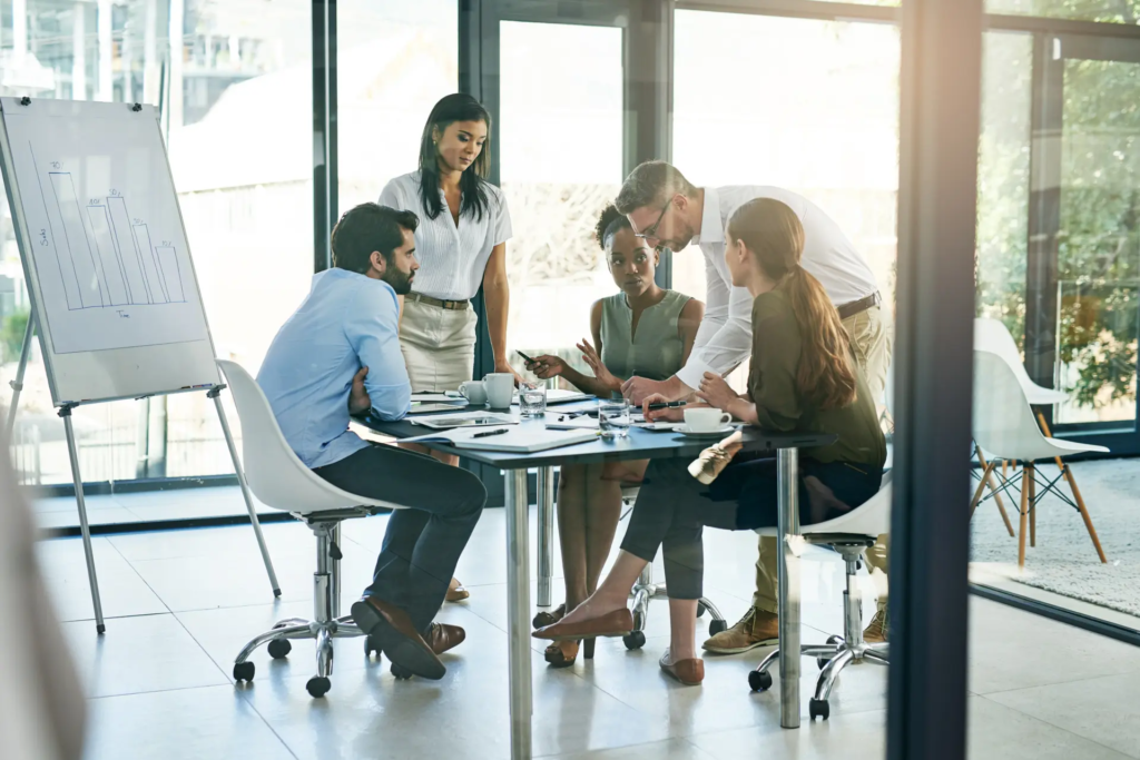 Un groupe de cinq professionnels est réuni autour d'une table de conférence en verre dans un bureau lumineux, discutant et collaborant. Un homme debout pointe l'écran d'un ordinateur portable tandis que les autres interagissent.