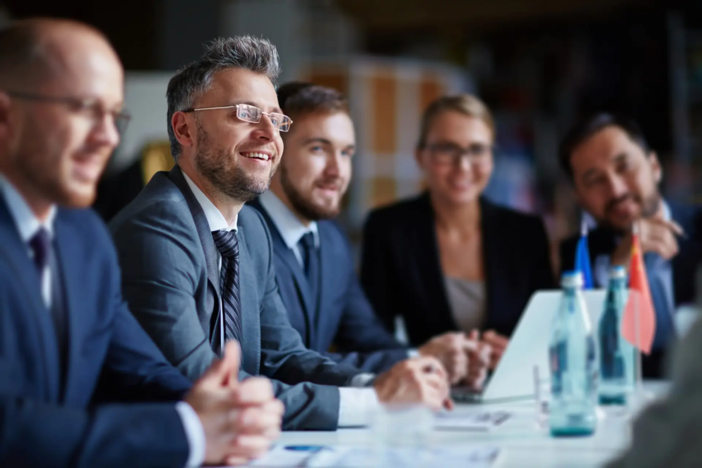 Un groupe de professionnels souriants et attentifs est assis autour d'une table de conférence, participant à une réunion. Des drapeaux miniatures sont visibles sur la table, suggérant une rencontre internationale ou diplomatique.