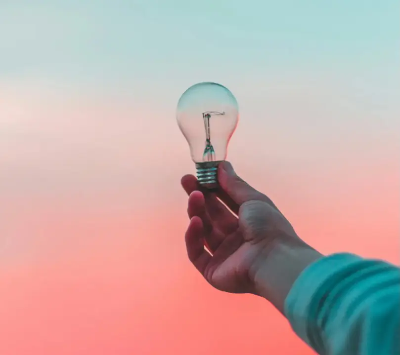Une main tient une ampoule classique, symbolisant une idée ou l'énergie, contre un ciel dégradé aux couleurs douces du crépuscule, passant du bleu clair au rose orangé.