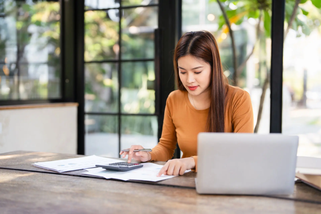 Une femme habillée d'un haut orange à son bureau. Elle lit des documents papier et utilise une calculatrice.