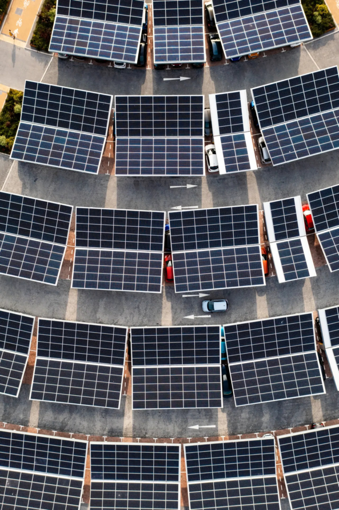 Vue aérienne d'un grand parking où les places de stationnement sont couvertes par des auvents de panneaux solaires. Quelques voitures sont visibles sous les structures, illustrant une infrastructure combinant stationnement et production d'énergie solaire.
