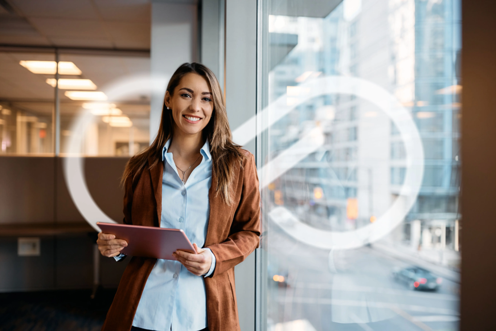 Femme debout près d’une fenêtre dans un bureau, souriante, tenant une tablette devant un espace de travail